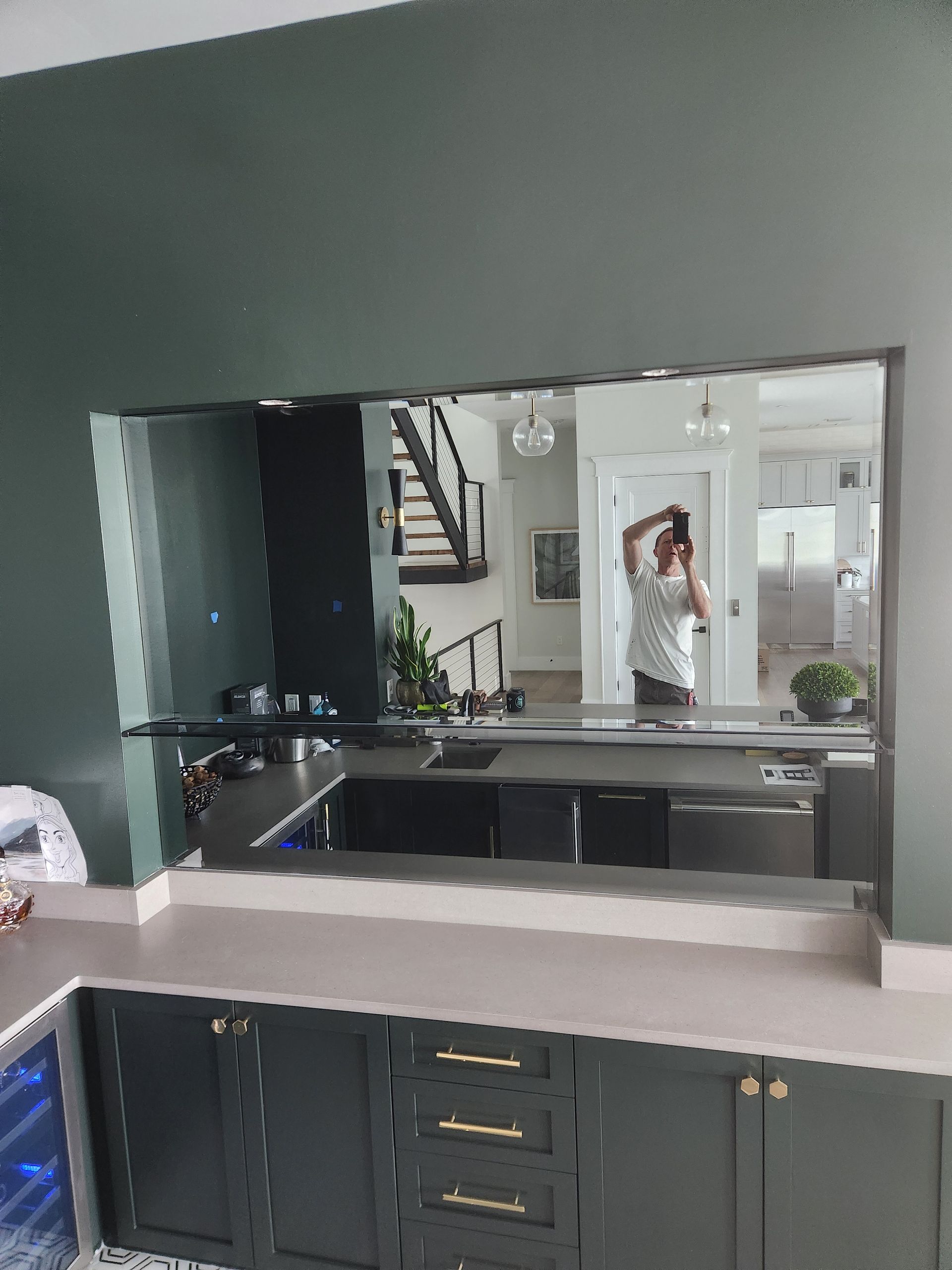 A modern kitchen pass-through with a mirror reflecting a person, stairs, and bright rooms; dark green cabinets.