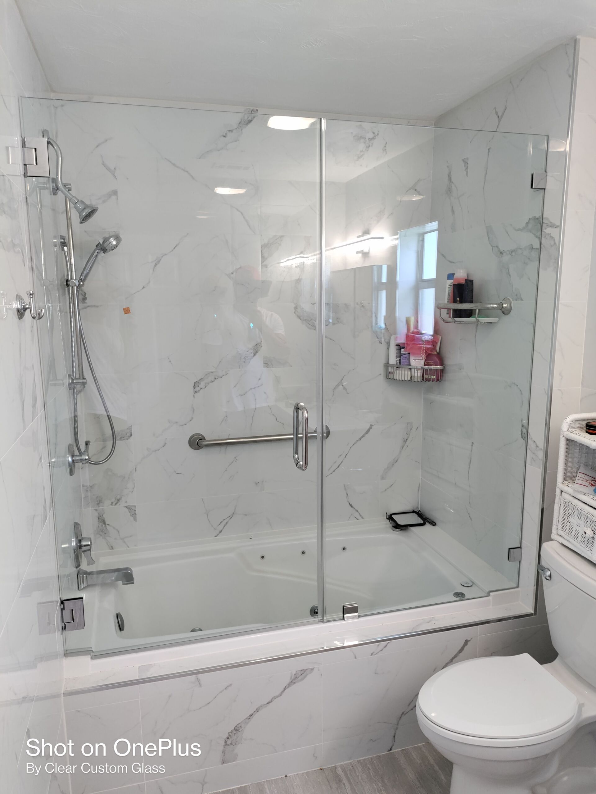 Glass-enclosed shower over a bathtub with marble-patterned walls, a toilet is in the foreground.