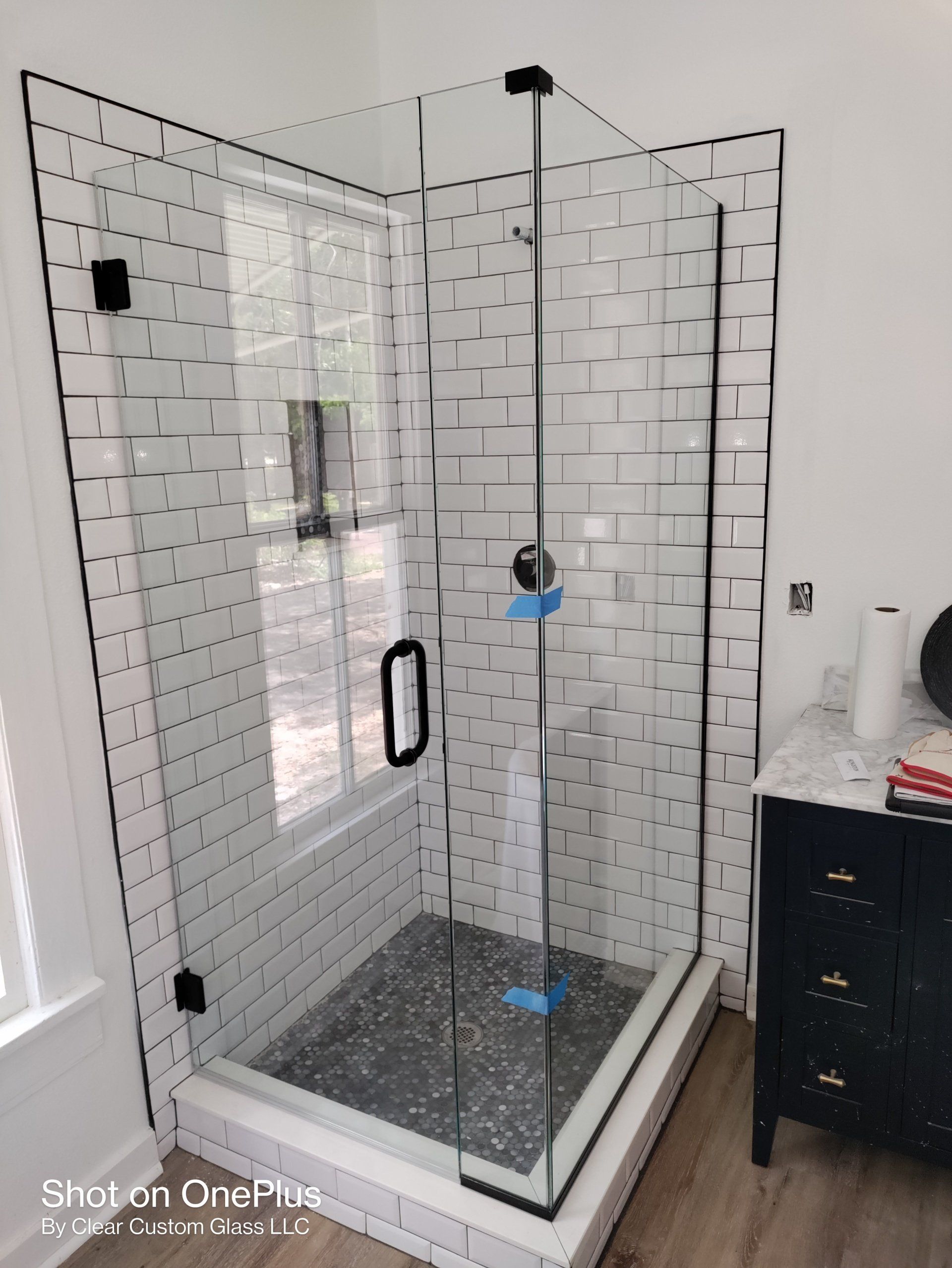 Corner shower with glass doors, black hardware, and subway tile walls; next to a window and cabinet.