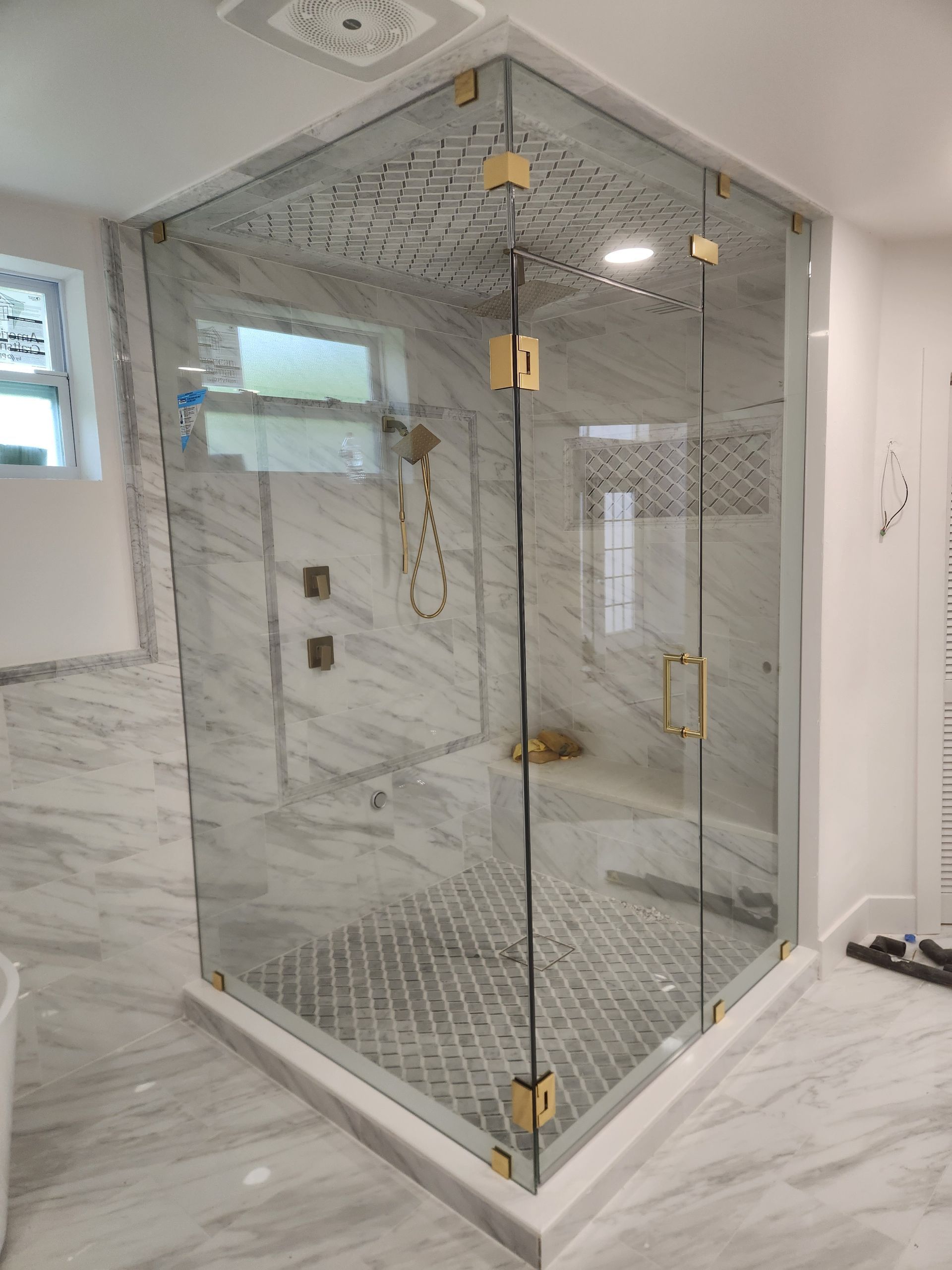 Corner shower with glass doors, gold fixtures, and white marble walls and flooring.