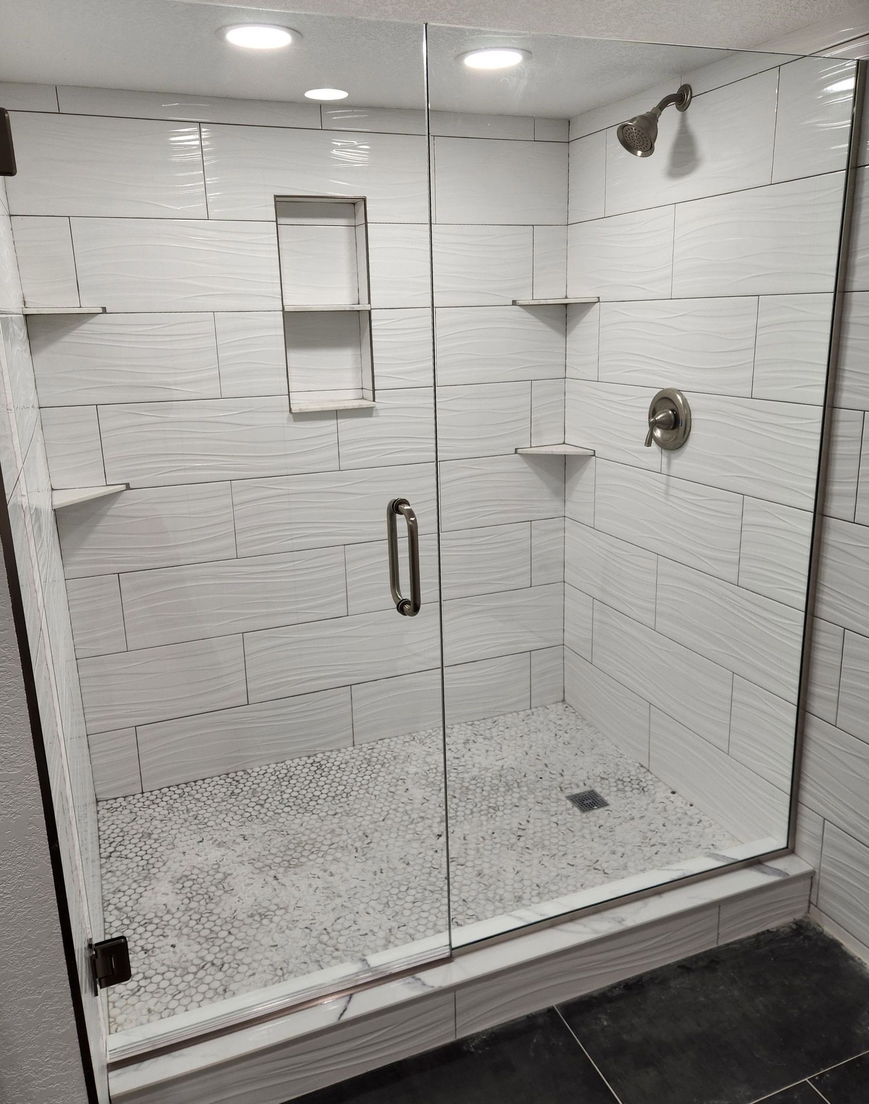 White-tiled shower with glass doors, built-in shelves, and a pebble-stone floor.
