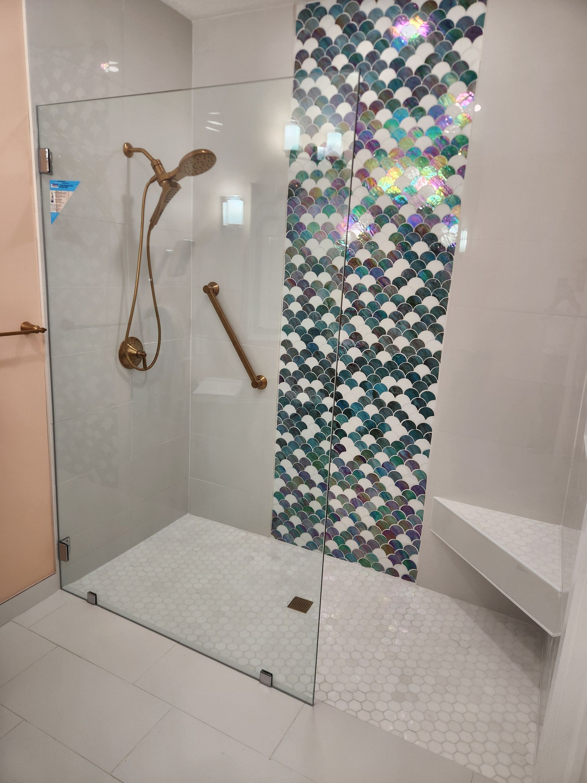 Modern shower with glass door, gold fixtures, mosaic accent wall, and corner seat.