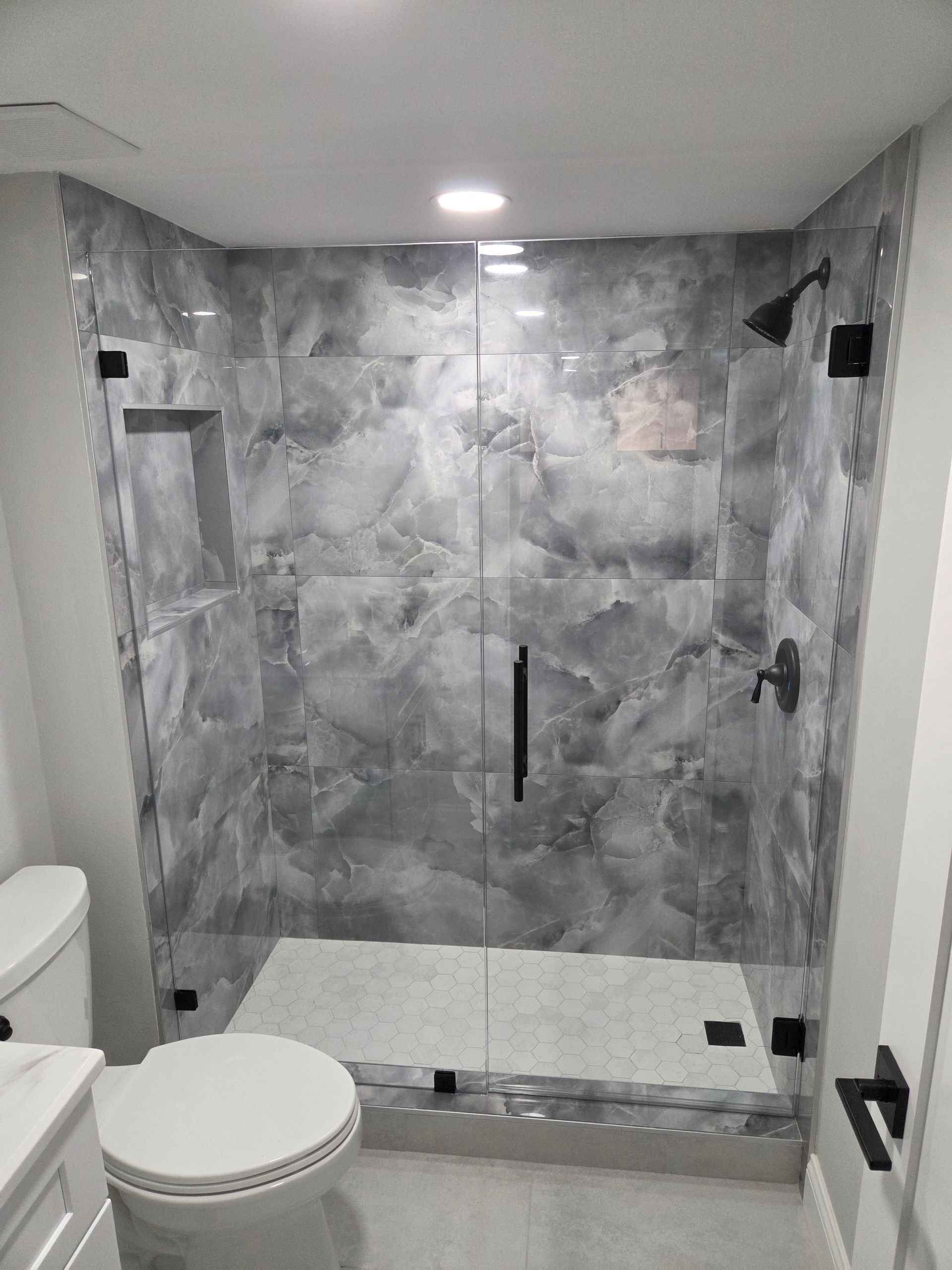 Modern shower enclosure with gray marble look tile, glass doors, and black fixtures. A toilet is visible to the left.