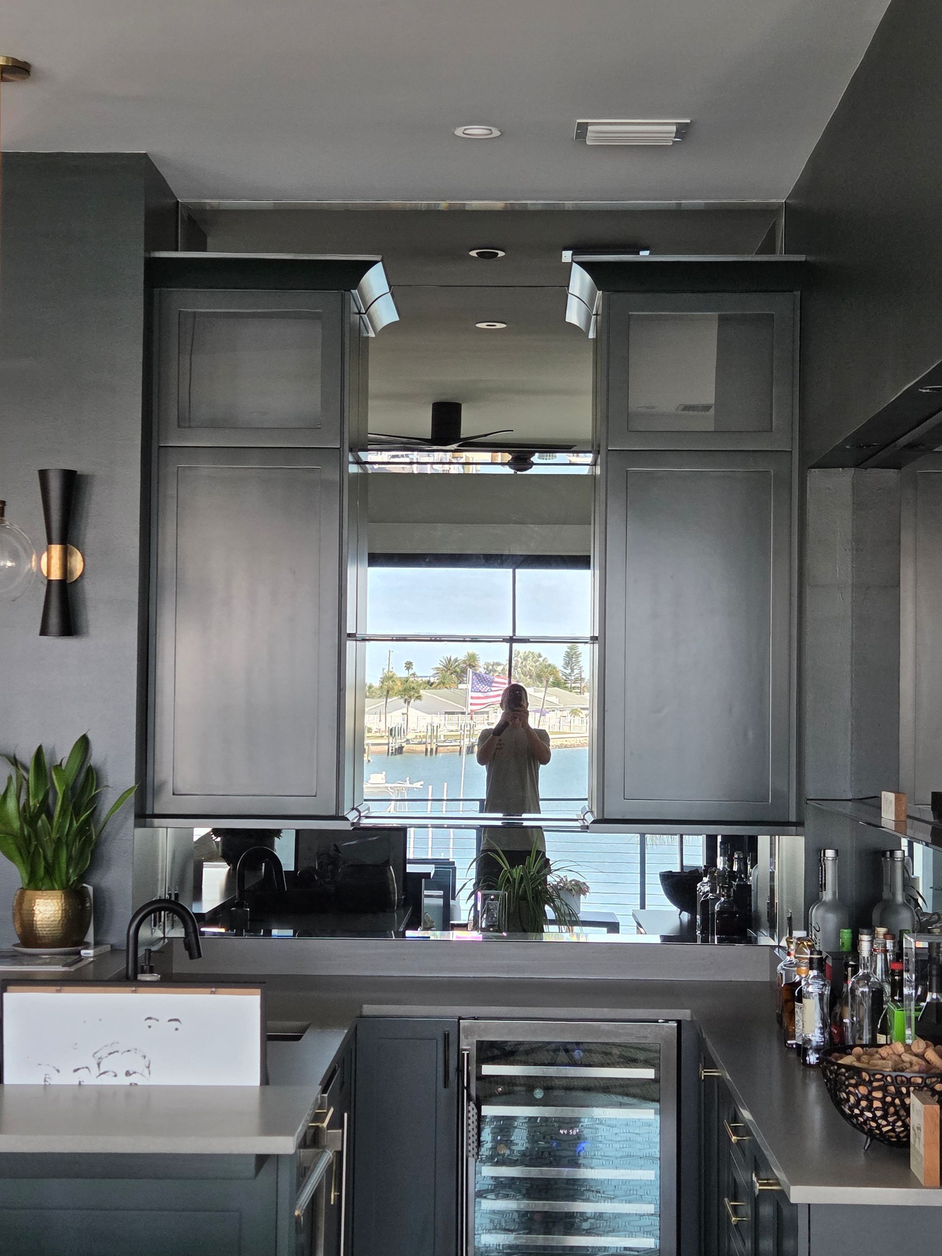 Kitchen with grey cabinets, mirrored upper section, and a person reflected in the window.