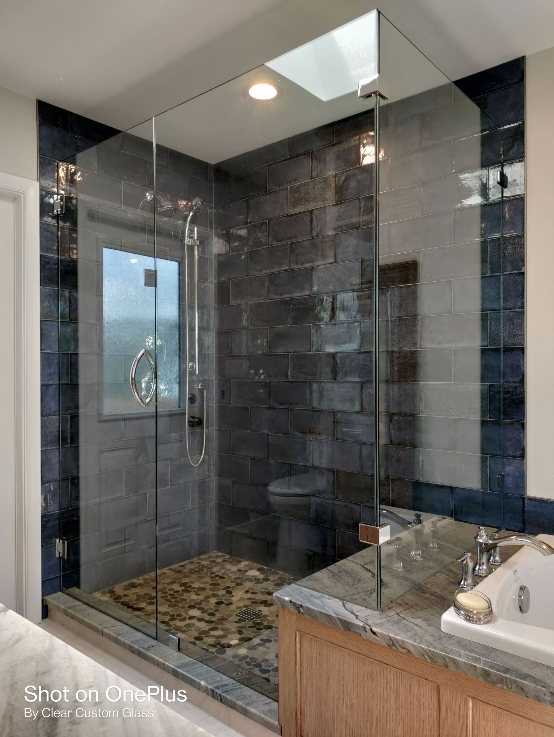 Modern shower with glass walls, stone floor, dark gray tile. Wood vanity and countertop.