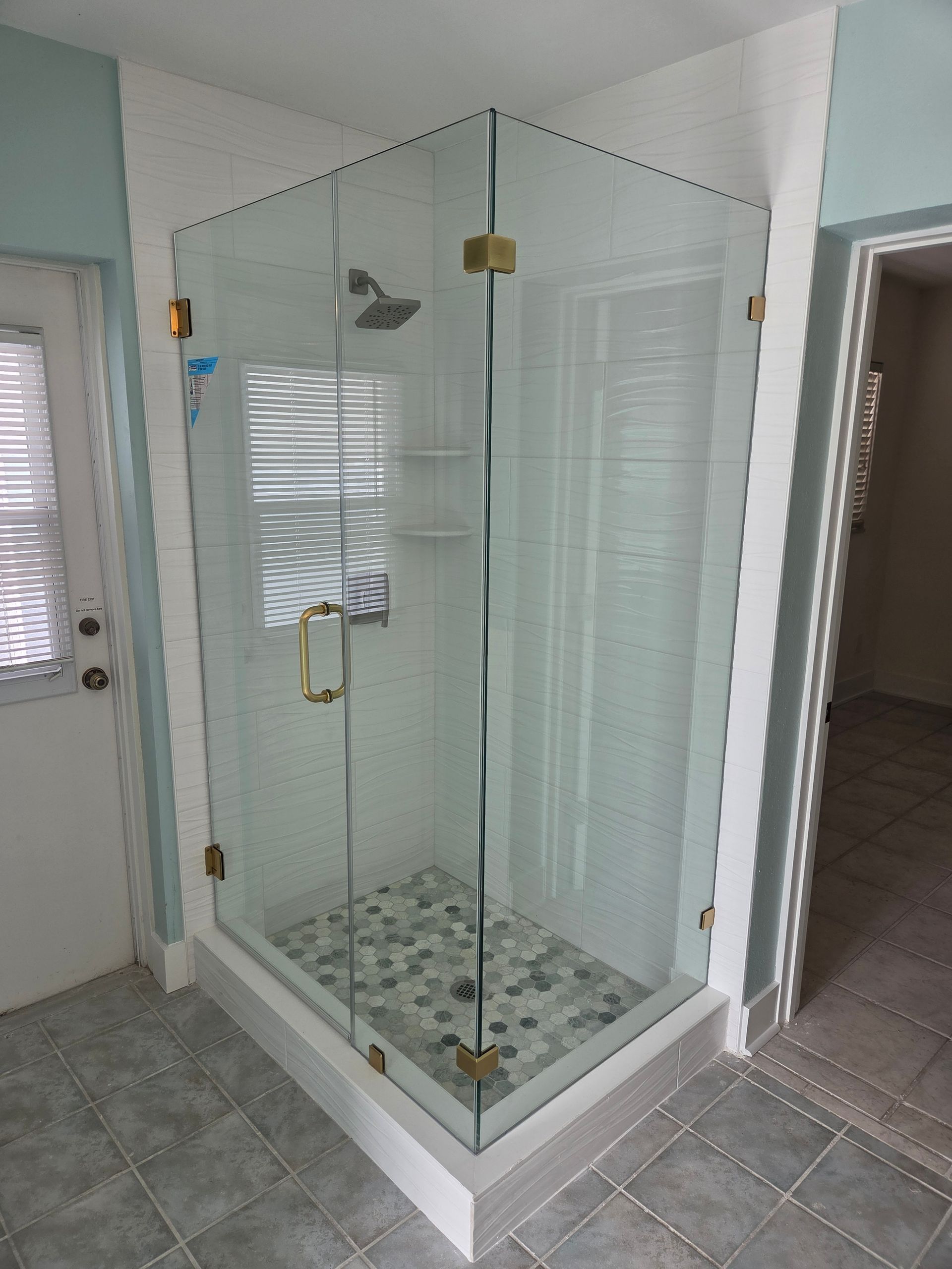 Glass-enclosed corner shower with tiled floor in a bathroom, beside a door and window blinds