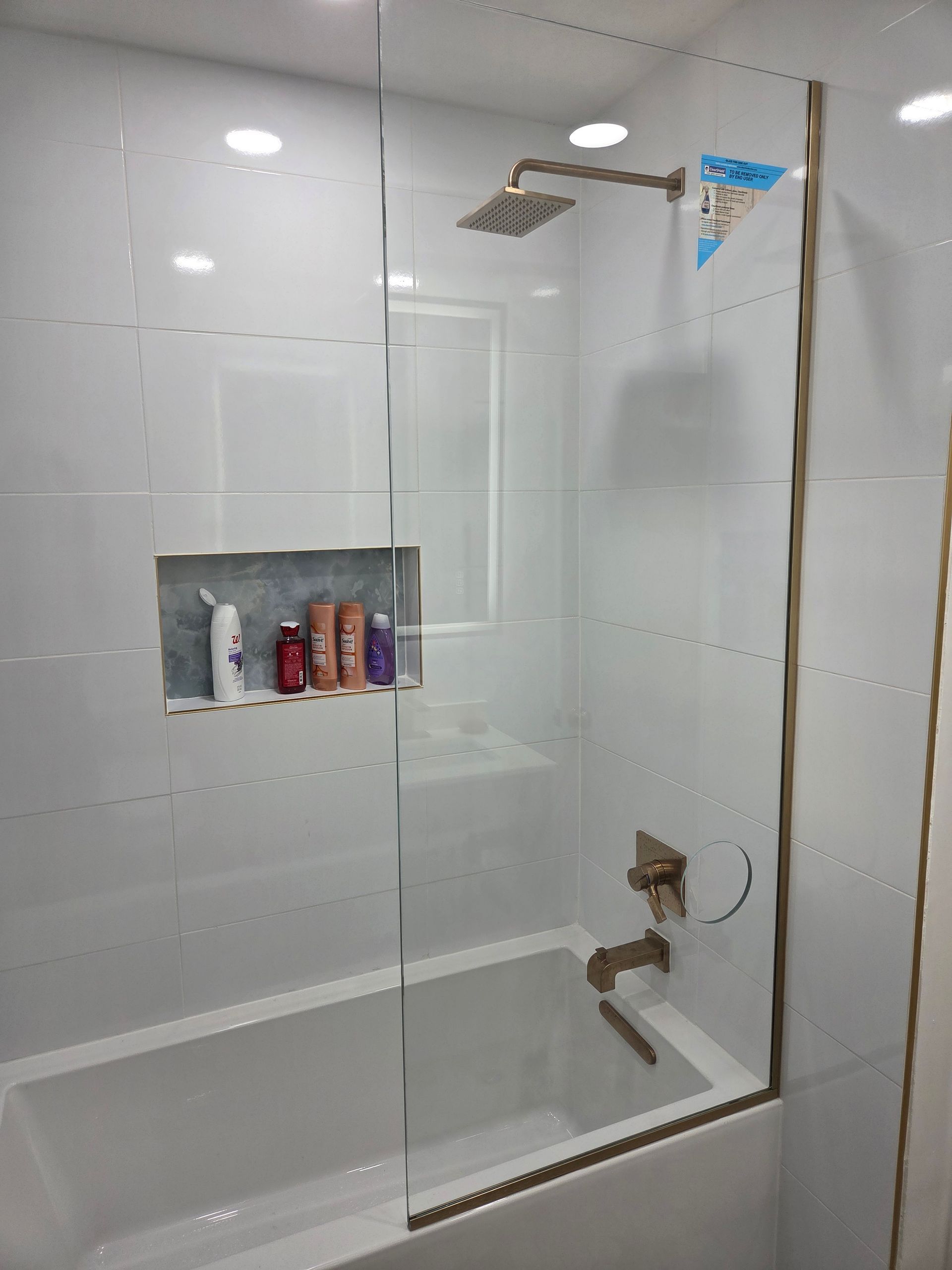 Bathroom with white tiled walls, glass shower door, gold fixtures, and a recessed shelf holding toiletries.
