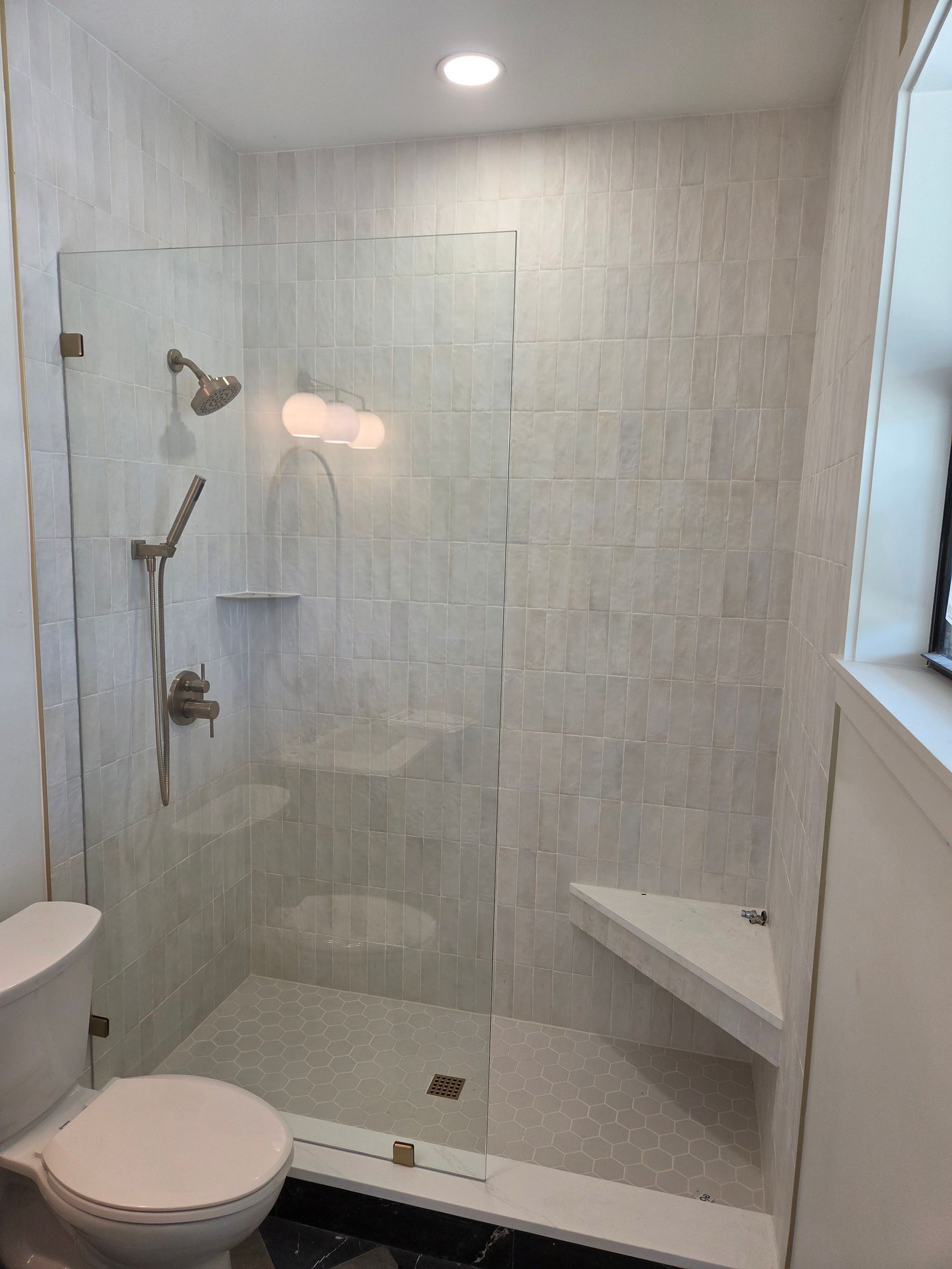 Bright tiled bathroom with glass shower, wall-mounted bench, toilet, and window