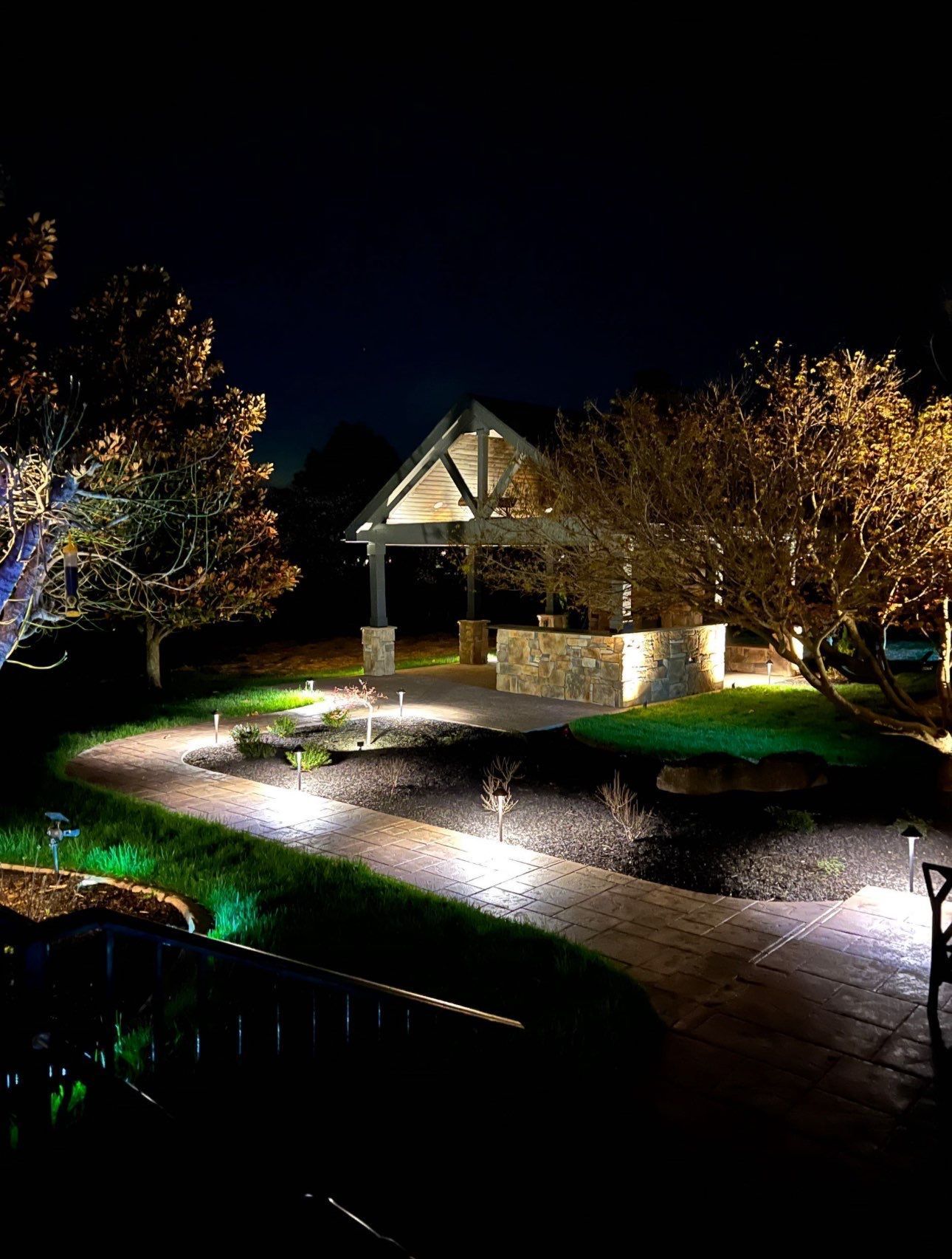 Illuminated brick house and front yard at night, with lit walkway and trees