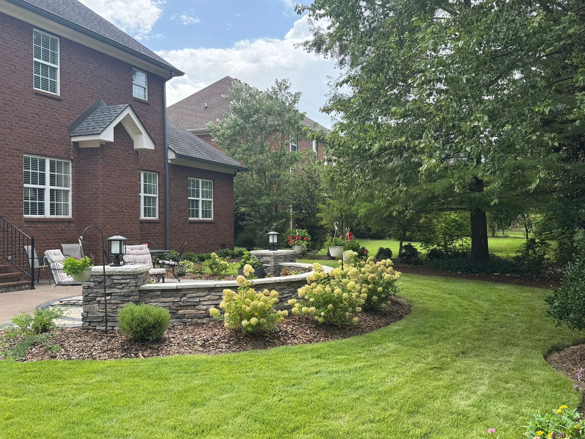 Brick house with landscaped garden, stone patio, and green lawn beside large trees