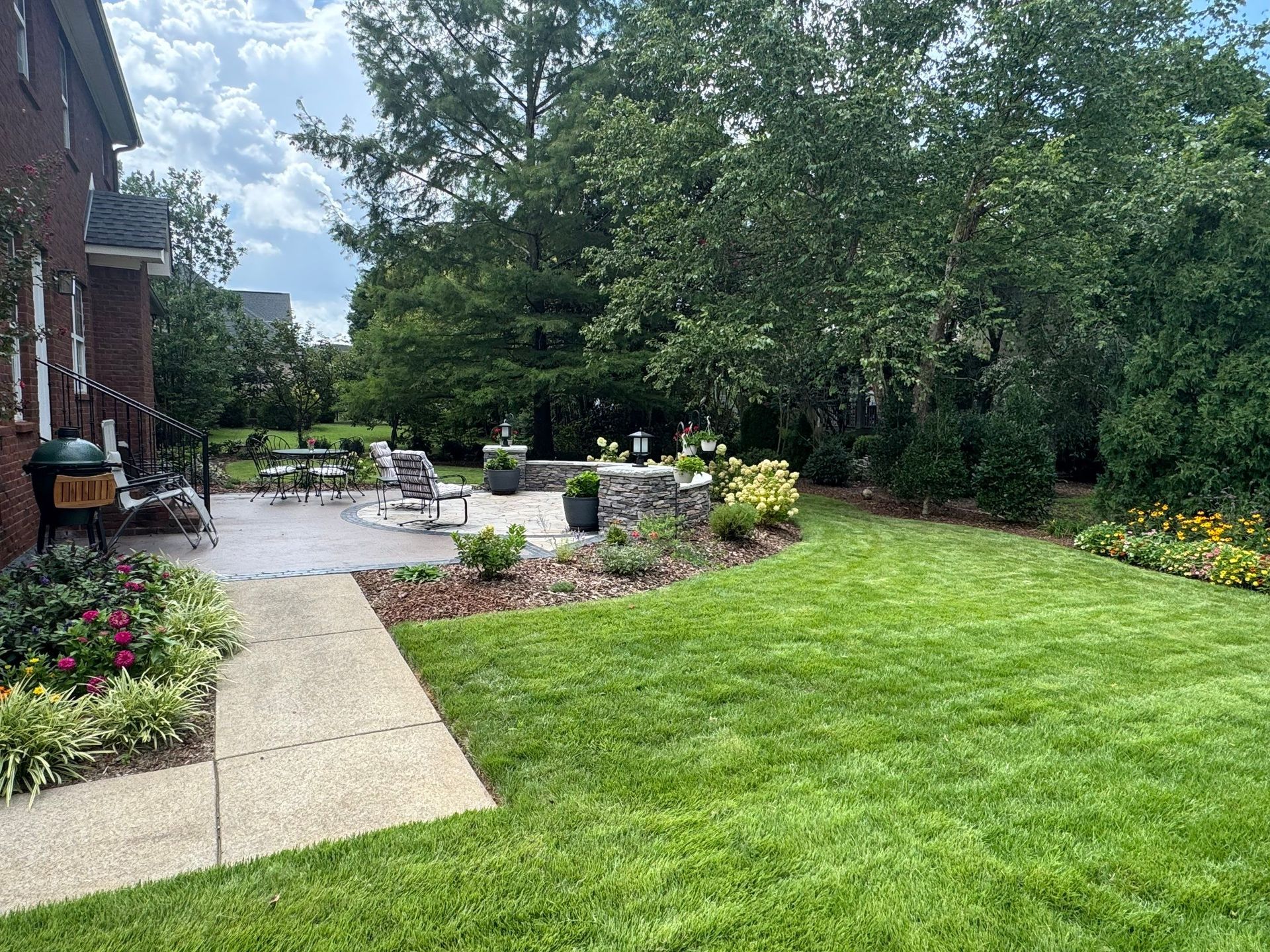 Backyard patio with stone walkway, green lawn, flowers, and trees beside a brick house