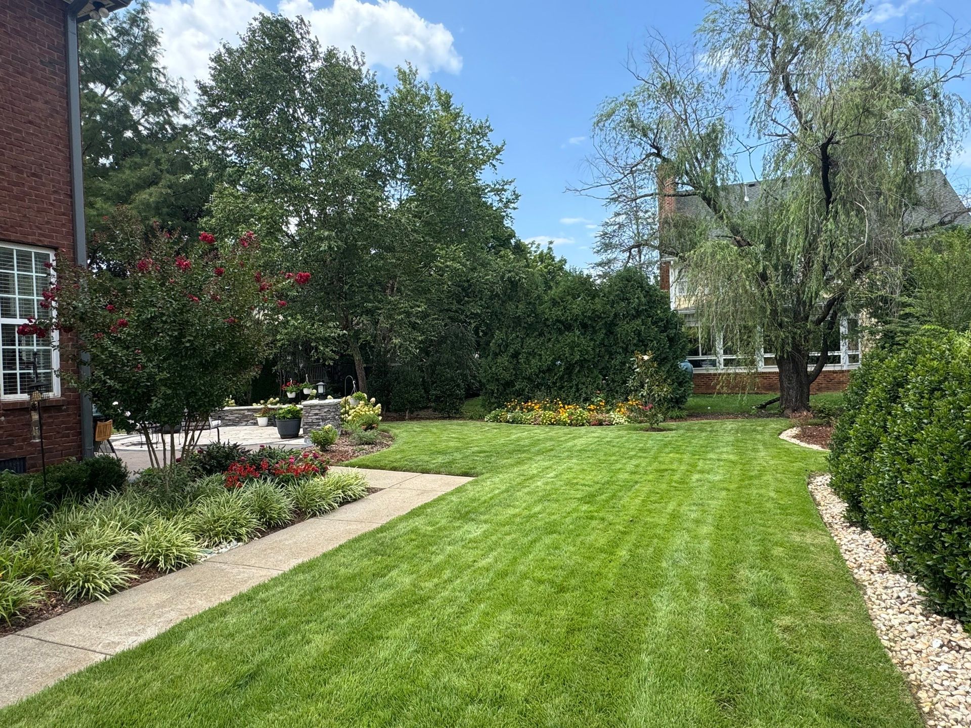 Lush backyard garden with green lawn, flowerbeds, trees, and a brick house on the left