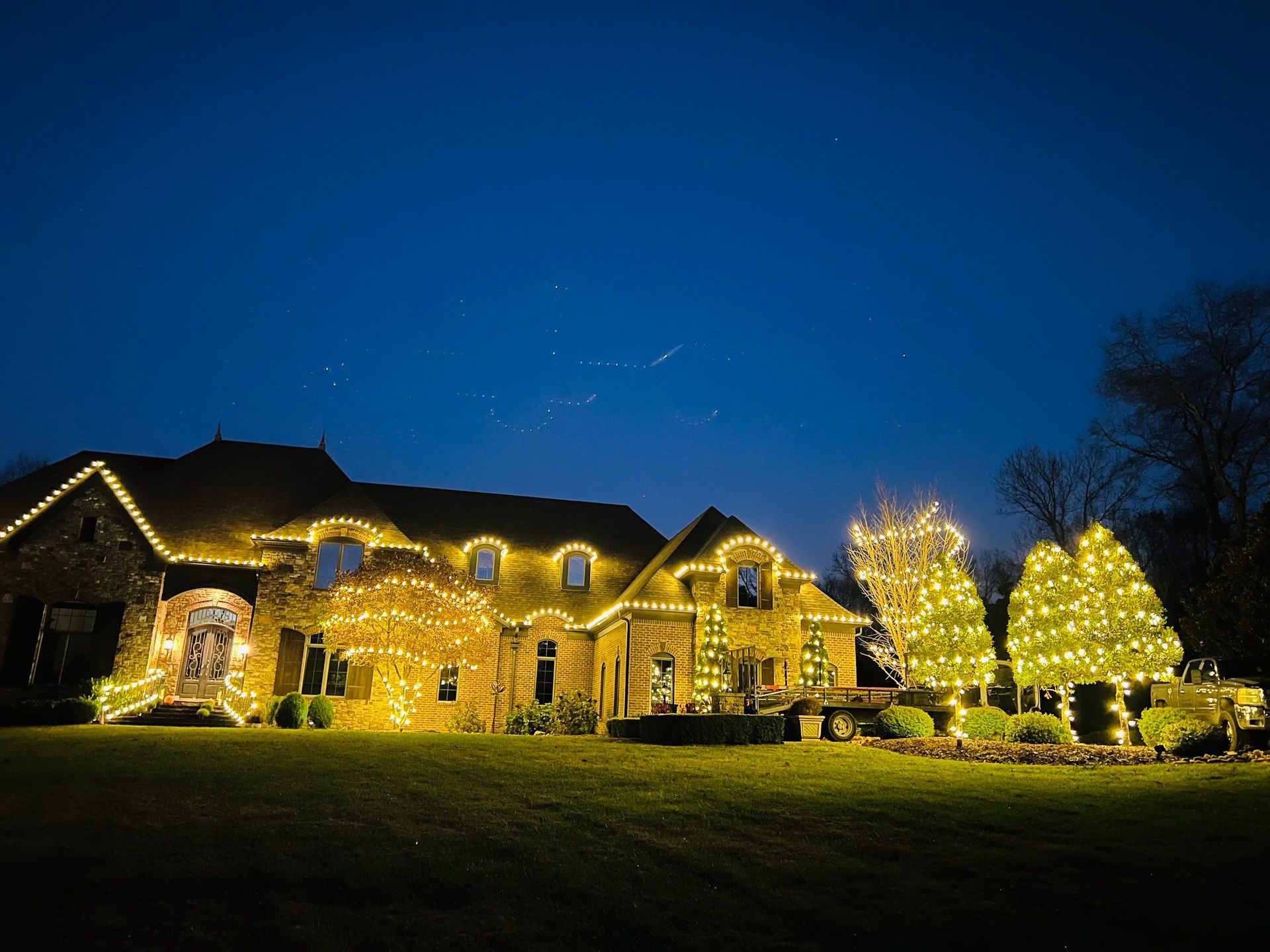 Large stone house at night, warmly lit with string lights and glowing windows under a deep blue sky.
