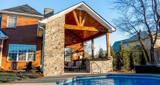Brick house with stone patio cover overlooking a pool. Brown wooden beams and ceiling.