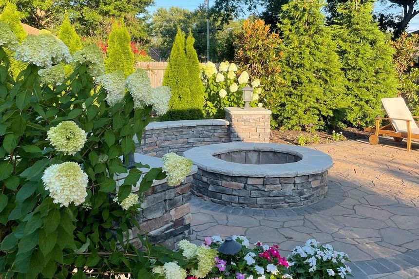 Stone fire pit and seating area in a backyard, with manicured plants and flowers.