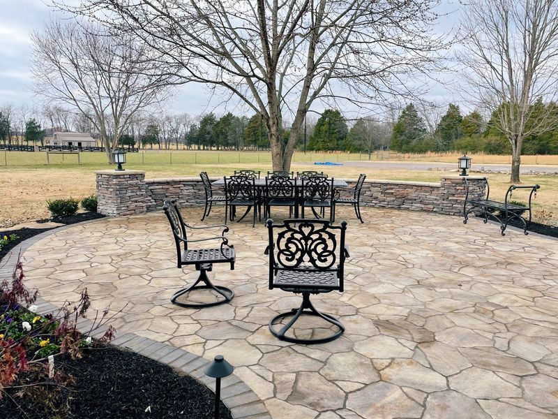Stone patio with wrought iron furniture, low stone wall, and trees on a cloudy day.