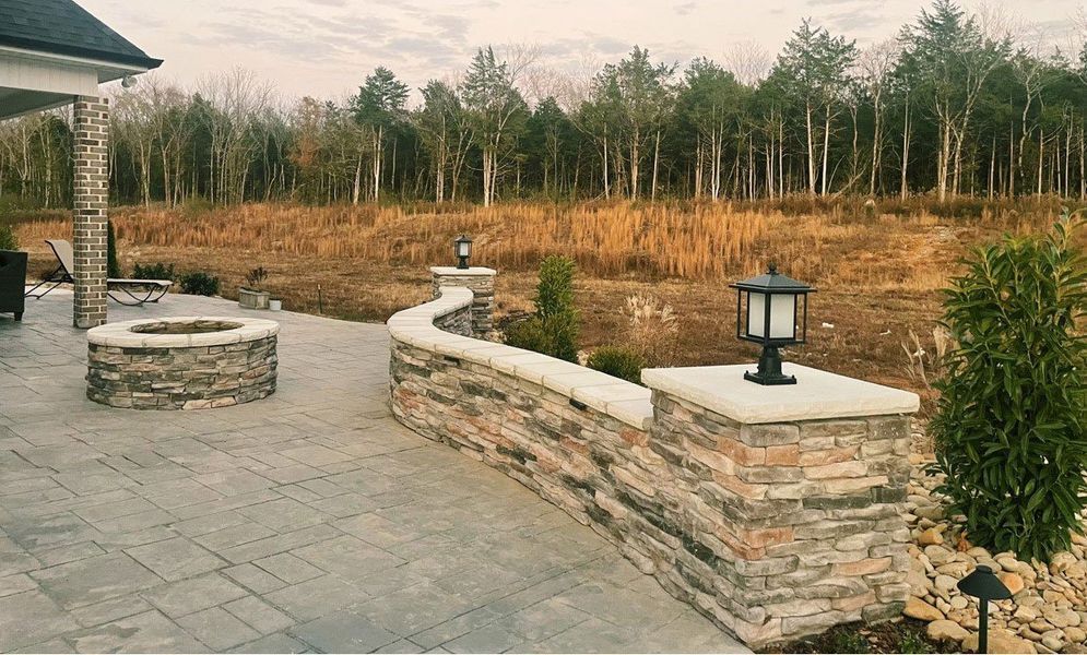 Stone patio with fire pit and low wall, overlooking a field with trees.