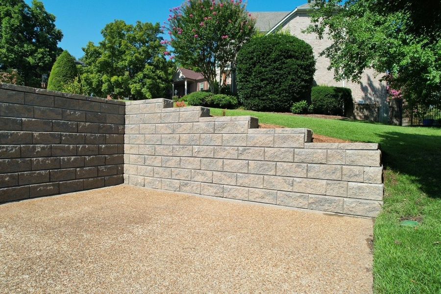 Stone retaining walls with stepped design and gravel driveway. House and green lawn in the background.