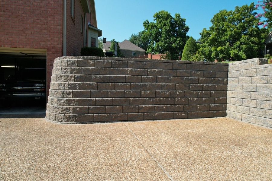 Retaining wall made of gray blocks beside a gravel driveway and a brick building.