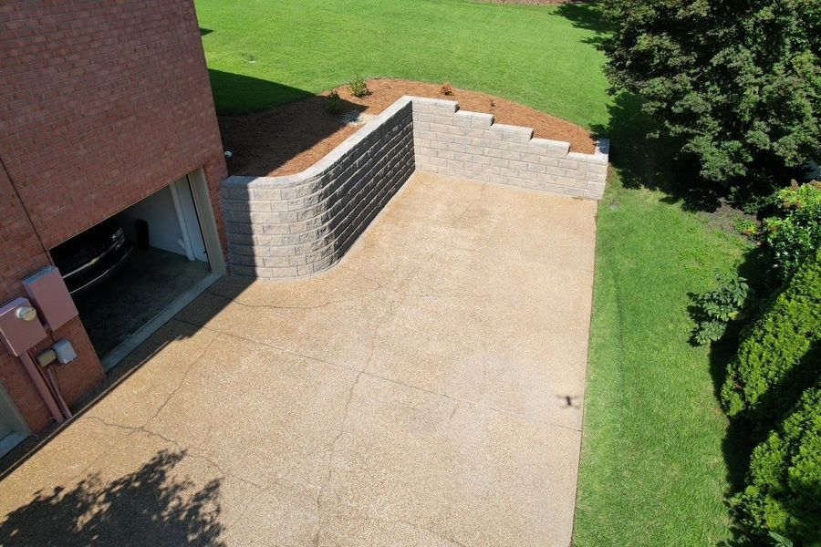 Concrete driveway next to a red brick building and a retaining wall with grass and trees.