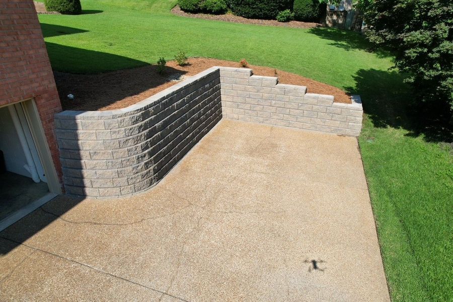 A retaining wall made of stone blocks surrounds a concrete patio next to a brick building with a grassy area behind.