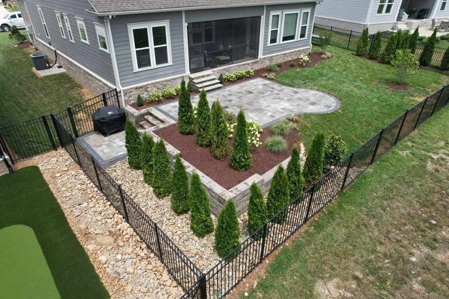 Backyard with a gray house, patio, stone path, black fence, green grass, and evergreen trees.