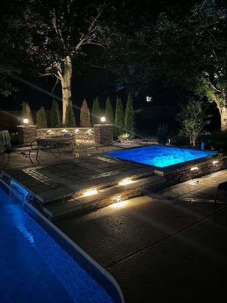 Nighttime backyard scene with a pool, spa, and landscape lighting. Blue water glows, stone features are lit.