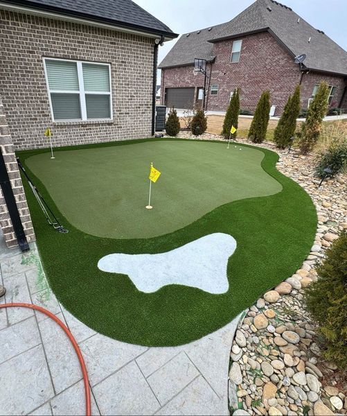 Backyard putting green with flags, sand trap, and landscaping; next to a brick house.