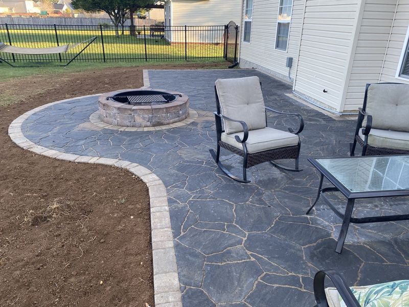 Stamped concrete patio with fire pit, seating, and beige trim, beside a light-colored house.