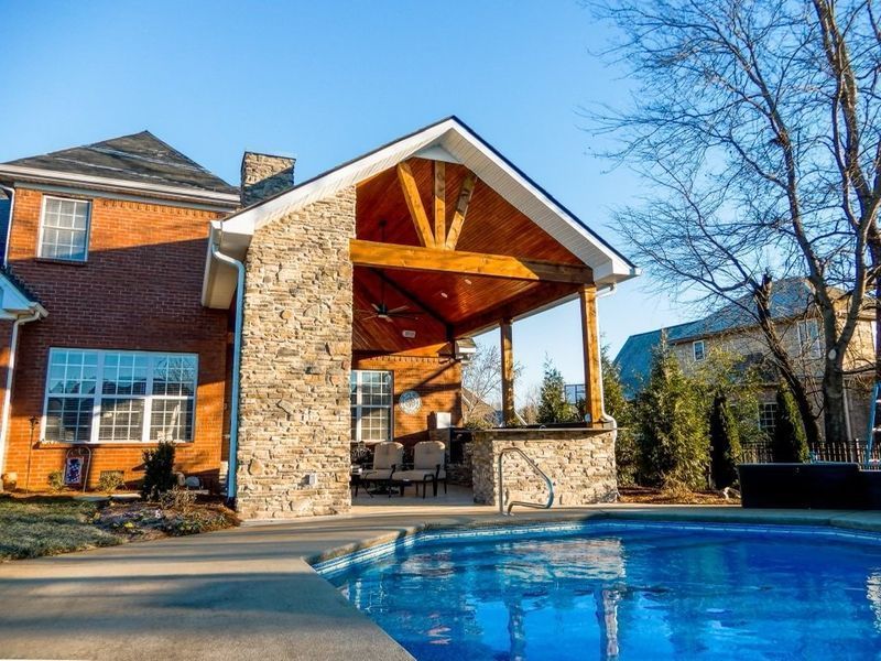 Backyard with a pool, covered patio, and brick house under a bright blue sky.