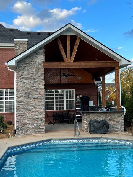 Poolside patio with stone facade, wooden beams, and a brick house, blue pool.