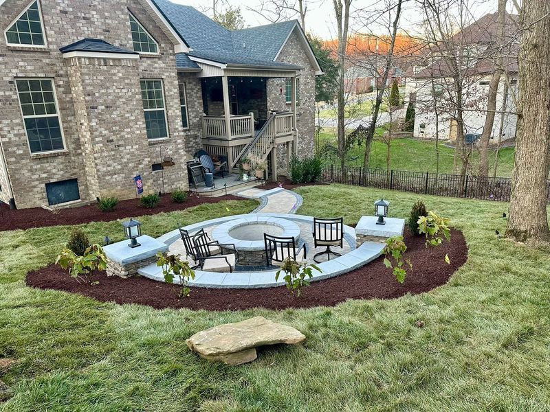 A backyard with a brick house, a fire pit area with chairs, and a stone bench on green grass.