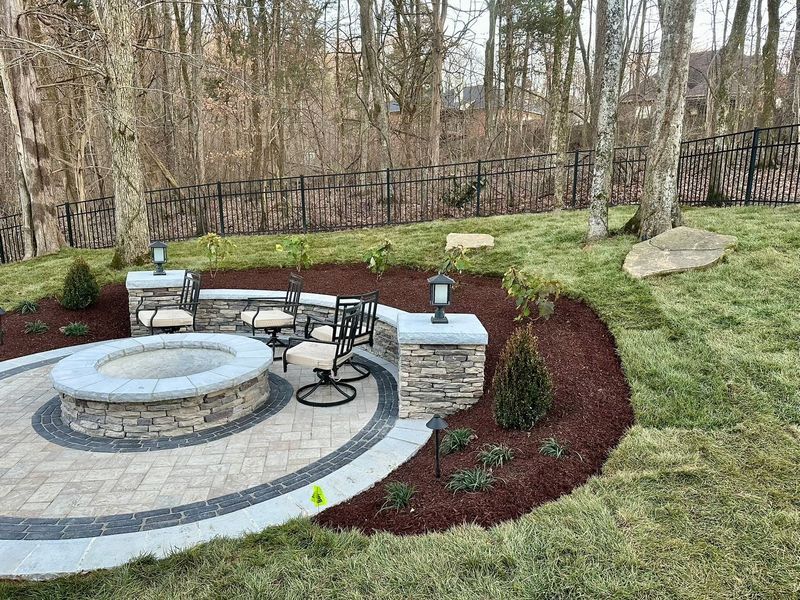 Patio with stone fire pit and seating area, surrounded by landscaping, trees, and a black fence.