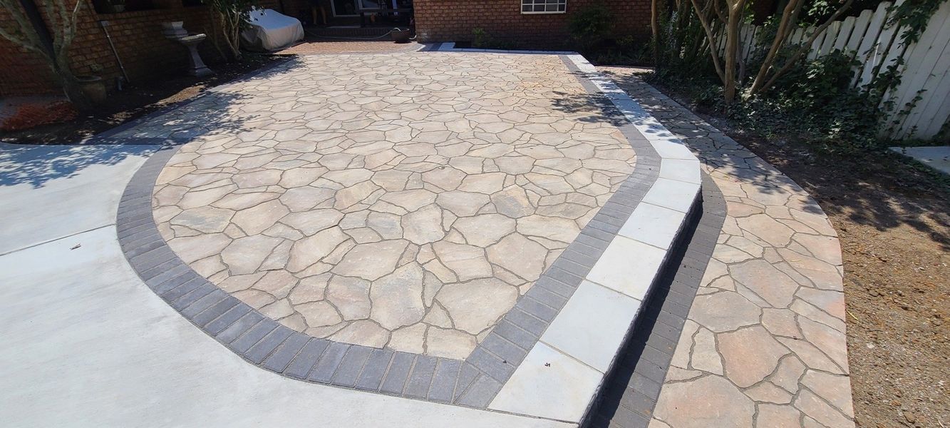 A driveway with a stone design, bordered by gray concrete and a paved walkway.