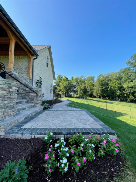 Stone patio with flower bed in front of a house, lawn and trees visible in the background under a blue sky.