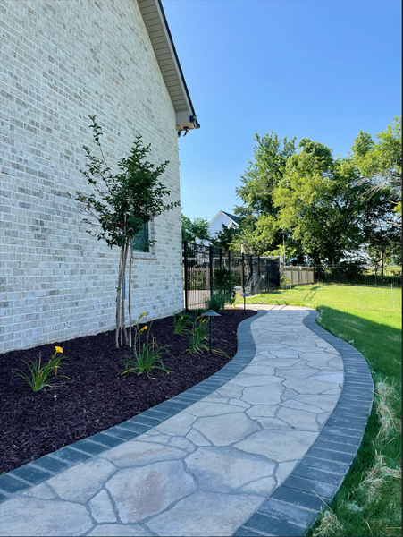 Stone path curves along brick wall, lined with plants and mulch.
