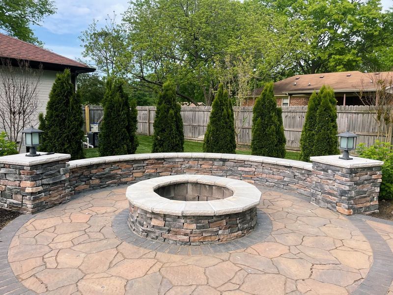 Stone patio with a fire pit and curved retaining wall, framed by evergreen trees, and surrounded by two houses.