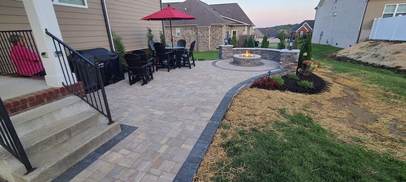 Patio with fire pit, seating area, and landscaping; red umbrella, gray pavers, and green grass.
