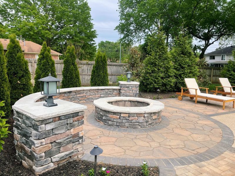Stone fire pit patio with seating, surrounded by evergreen trees and a wooden fence.