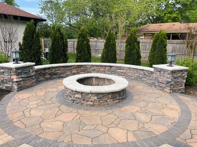 A fire pit area with stone pavers, a curved wall, and evergreen trees in a backyard.
