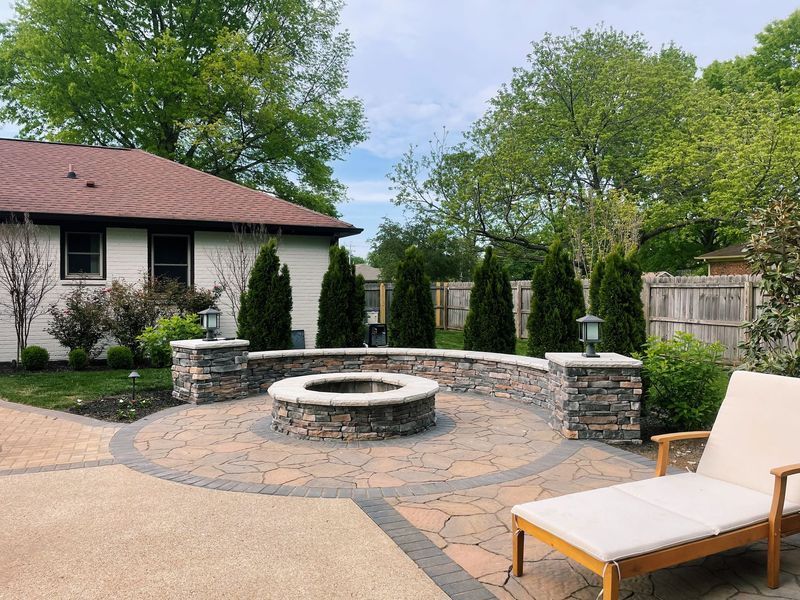 A stone patio with fire pit, surrounded by pillars with lanterns and evergreen trees.