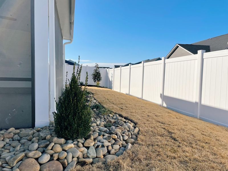 Backyard with white fence, dry grass, rocks, and a small evergreen bush under a bright blue sky.