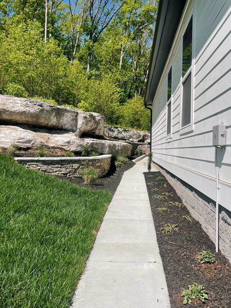 Concrete walkway alongside a white house with a rock retaining wall and green grass.