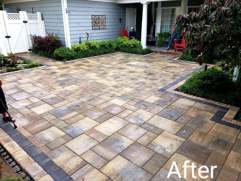 Paver patio with border next to a house, with greenery and a gate. 