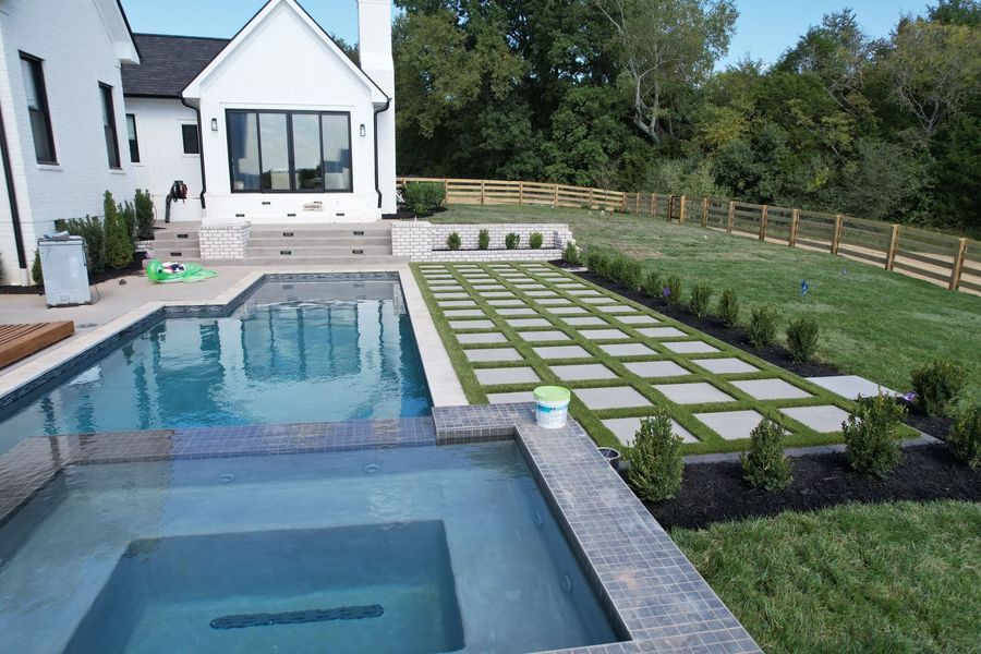 Backyard pool with square pavers and green grass, next to a white house and wooden fence.