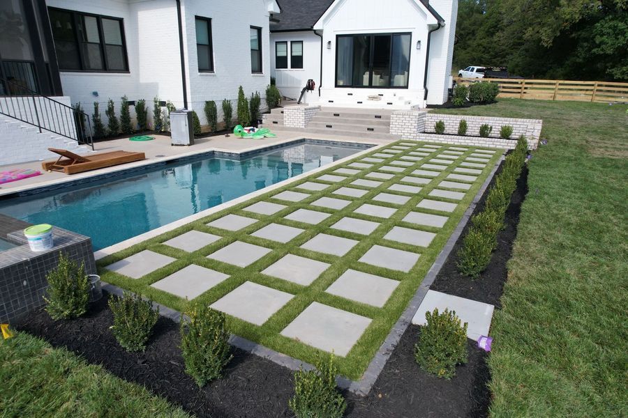 A rectangular pool with a tiled walkway, surrounded by manicured grass and shrubs, near a white house.