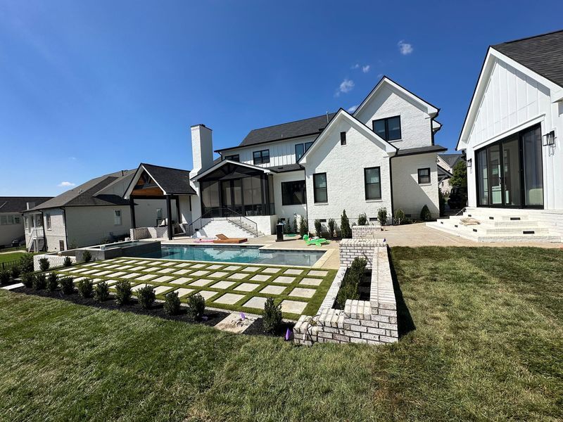 Backyard with pool, white brick house, blue sky. Lawn, stone pavers, and greenery surround the pool area.