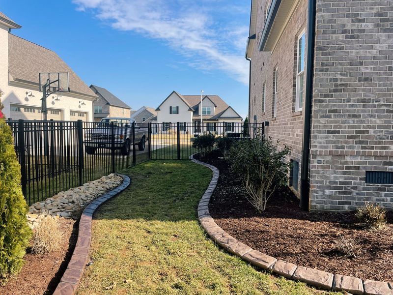 A residential backyard with a brick house, green grass, black fence, and sunny sky.