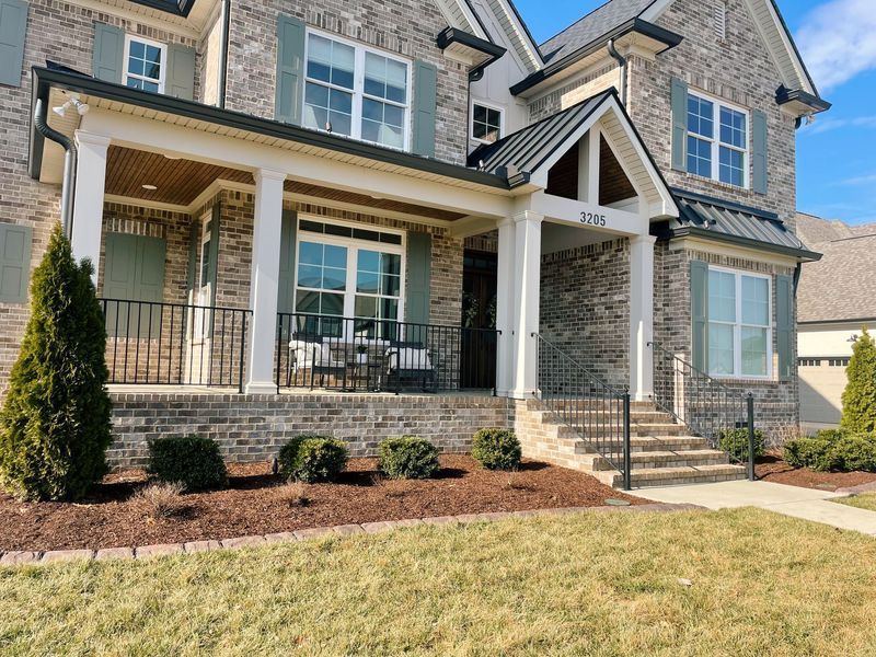 Two-story brick house with a porch, steps, shutters, and a manicured lawn under a sunny sky.