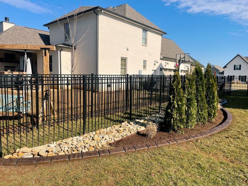 Black metal fence encloses a backyard with trees, a brick house, and a stamped concrete border.